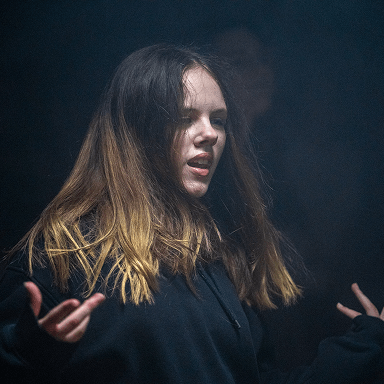 Junge Frau mit langen braun-blonden Haaren und schwarzem Kapuzenpullover vor dunklem Hintergrund