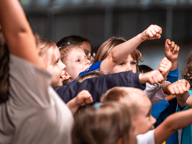 Kinder in bunter Kleidung strecken synchron die Arme nach vorne in einem Innenraum