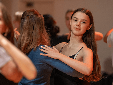 Zwei junge Frauen im Tanzkurs in elegantem Abendkleid und blauer Bluse in hellem Tanzsaal mit weiteren Paaren im Hintergrund